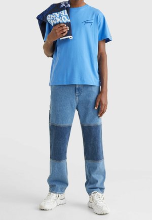 Henkilö, joka pukeutuu siniseen t-paitaan, tilkkutäkkihousuihin, valkoisiin lenkkareihin ja pitää navy-väristä Tommy Jeans -neuletta toisen olkapään yli.