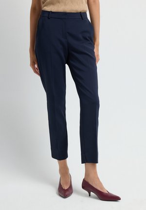 Woman wearing navy blue tailored ankle-length pants and burgundy low-heeled pointed shoes, standing against a plain light background.