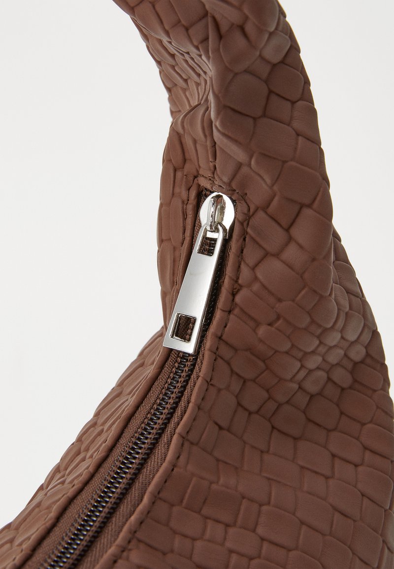 Sac à main texturé marron avec un motif reptilien, comportant une fermeture éclair et une tirette en argent. La poignée est incurvée et douce au toucher.