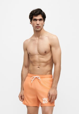 Jeune homme aux cheveux foncés portant un short de bain orange Hugo avec cordon blanc, se tenant devant un fond clair uni.