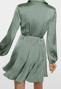 Robe en satin vert avec des manches longues, un col et une ceinture à nouer. La jupe s'évase avec des plis, mettant en valeur une texture lisse et un fluide élégant.