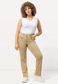 Beige raka byxor med dragkedjefickor, matchat med en vit ärmlös topp. Skorna är vita och har en texturerad design.