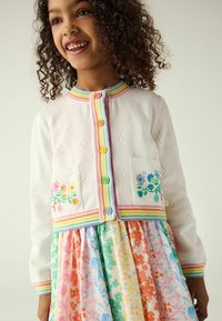 Cardigan bianco con motivo a occhielli, collo a strisce arcobaleno e ricami floreali colorati, abbinato a un vestito floreale multicolore.