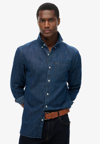 Hombre vestido con una camisa de mezclilla azul oscuro de botones, con el cuello levantado, manos detrás de la espalda, combinada con un cinturón de cuero marrón y jeans oscuros.