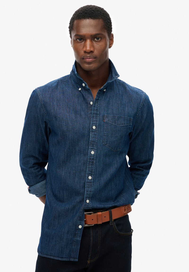 Hombre vestido con una camisa de mezclilla azul oscuro de botones, con el cuello levantado, manos detrás de la espalda, combinada con un cinturón de cuero marrón y jeans oscuros.