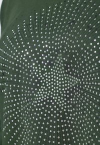 Chemise verte ornée d'un motif de strass argentés disposés en un design radieux, avec des lignes noires rehaussant la texture.