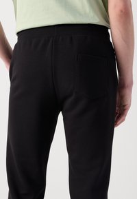 Pantaloni sportivi neri con un cinturino a coste in vita, dotati di una piccola tasca sul retro. Il tessuto appare liscio con una sottile trama.