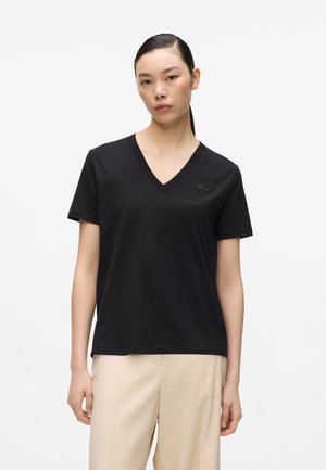 Ung kvinde med glat sort hår iført en sort T-shirt med V-hals og korte ærmer samt beige bukser, stående foran en ensfarvet hvid baggrund.