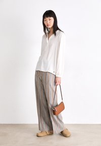 Blouse crème avec un col en V, manches longues ; pantalons larges rayés en beige, bleu et marron ; sac bandoulière marron ; sabots tan.