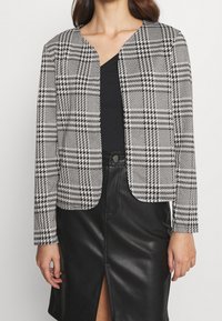 Blazer de patrón pata de gallo en blanco y negro con corte corto, diseño sin cuello y mangas largas, combinado con una blusa negra y una falda de cuero.