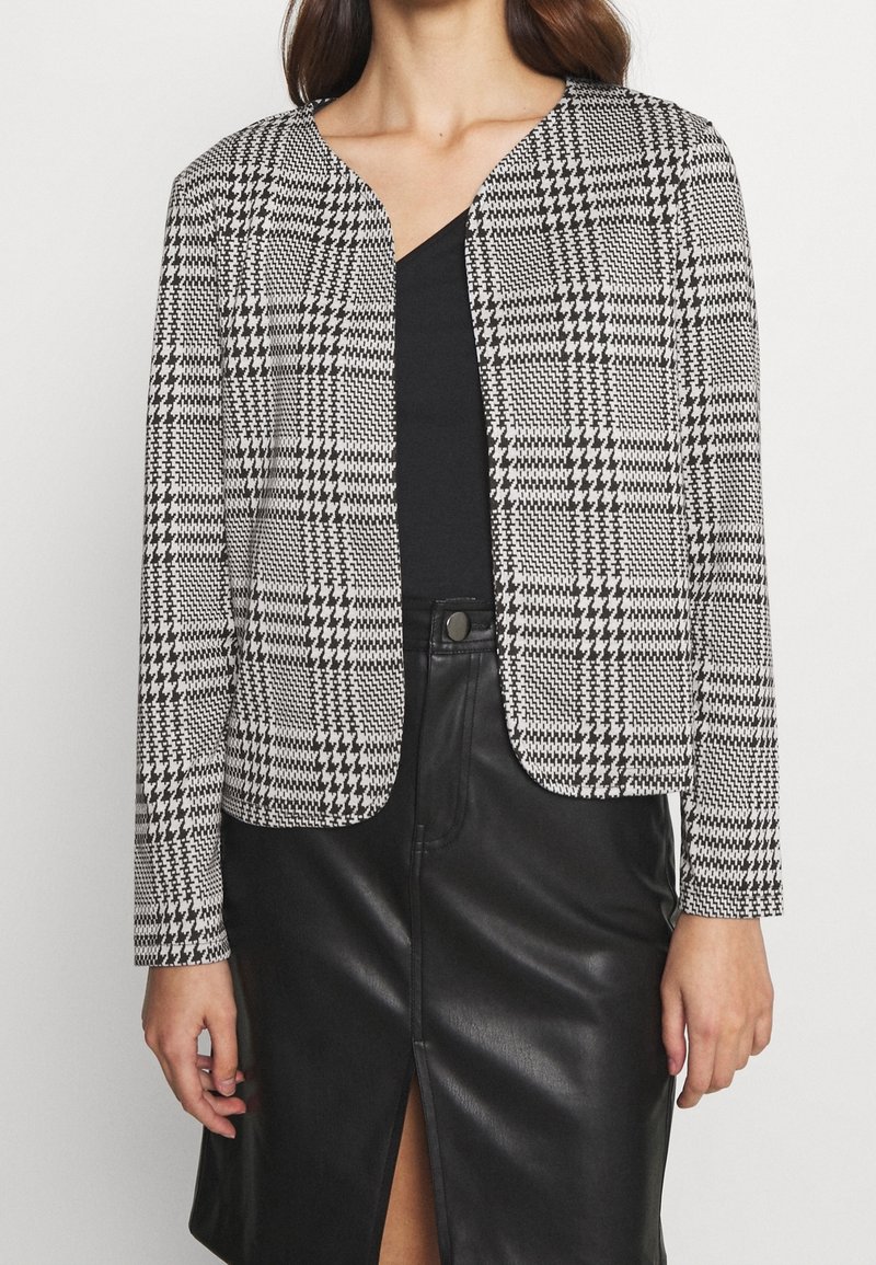 Blazer de patrón pata de gallo en blanco y negro con corte corto, diseño sin cuello y mangas largas, combinado con una blusa negra y una falda de cuero.