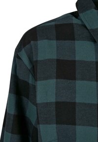 Gros plan sur l'épaule et le col d'une chemise en tissu à carreaux verts et noirs avec une texture douce et tissée.