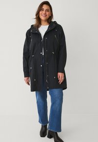 Zwarte hoodiejas met ritszakken, verstelbare trekkoord in de taille en metalen hardware-details, gecombineerd met blauw wijduitlopende jeans.
