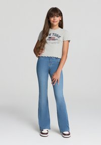 T-shirt grigia corta con bordi frangiati e testo "NEW YORK", abbinata a jeans flare blu a vita alta e sneakers bianche con dettagli bordeaux.