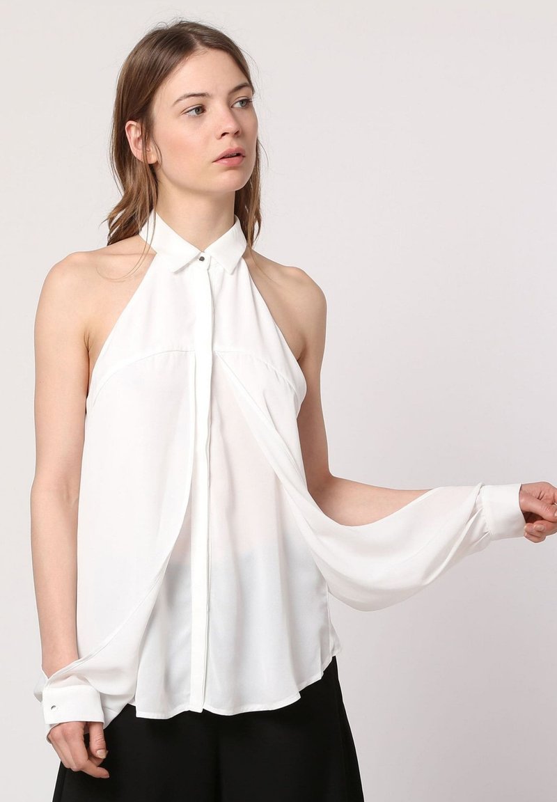 Blusa blanca sin mangas con cuello alto y tejido fluido. Presenta cierre de botones, mangas sueltas y un dobladillo curvado.