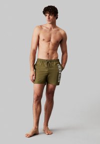 Pantaloni da nuoto verde oliva con vita elastica, chiusura con cordoncino e marchio bianco "CALVIN KLEIN" sul lato sinistro.