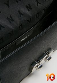 Černá kabelka s hladkým povrchem, kovovým zapínáním a vzorovanou podšívkou s logem "DKNY". Akcenty zahrnují lesklé kování.