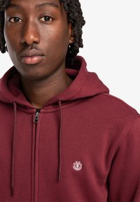 Felpa con zip di colore marrone scuro realizzata in tessuto morbido, con tasca a marsupio e un piccolo logo ricamato sul lato sinistro del petto.