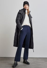 Kvinna i svart stickad mössa, lång svart pufferjacka, vit tröja, marinblåa byxor och svarta ankelstövlar som står på grått golv mot en neutral vägg.