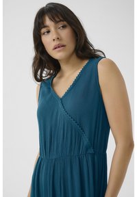 Teal dress made of soft, pleated fabric with a V-neck and scalloped trim. Features a fitted bodice and flowing skirt for a relaxed fit.