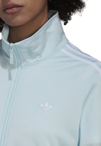 Primer plano de una persona que lleva una chaqueta de entrenamiento Adidas de color azul claro con un cuello alto, cremallera delantera y el logo de Adidas en blanco.