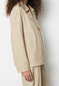 Beżowa bawełniana marynarka z kołnierzem z wcięciem, pojedynczym guzikiem i boczną kieszenią, o luźnym kroju i z minimalnymi detalami szwów.