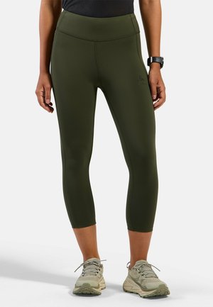 Person wearing olive green cropped leggings, beige running shoes, and a black smartwatch against a plain light background.