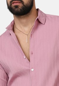 Maglietta rosa chiaro a coste con scollo a V, dotata di bottoni frontali, maniche corte e una collana con pendente argento con un accento dritto.