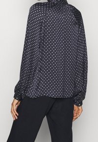 Blusa nera a fantasia con un design fluido, maniche lunghe con polsini arricciati e piccole forme di diamante di un colore più chiaro.