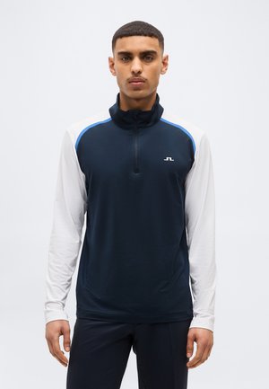 Mees, kes kannab tumesinist ja valget pika varrukaga spordijopet, millel on rinnal väike valge logo, seisab ühevärvilise tausta ees.