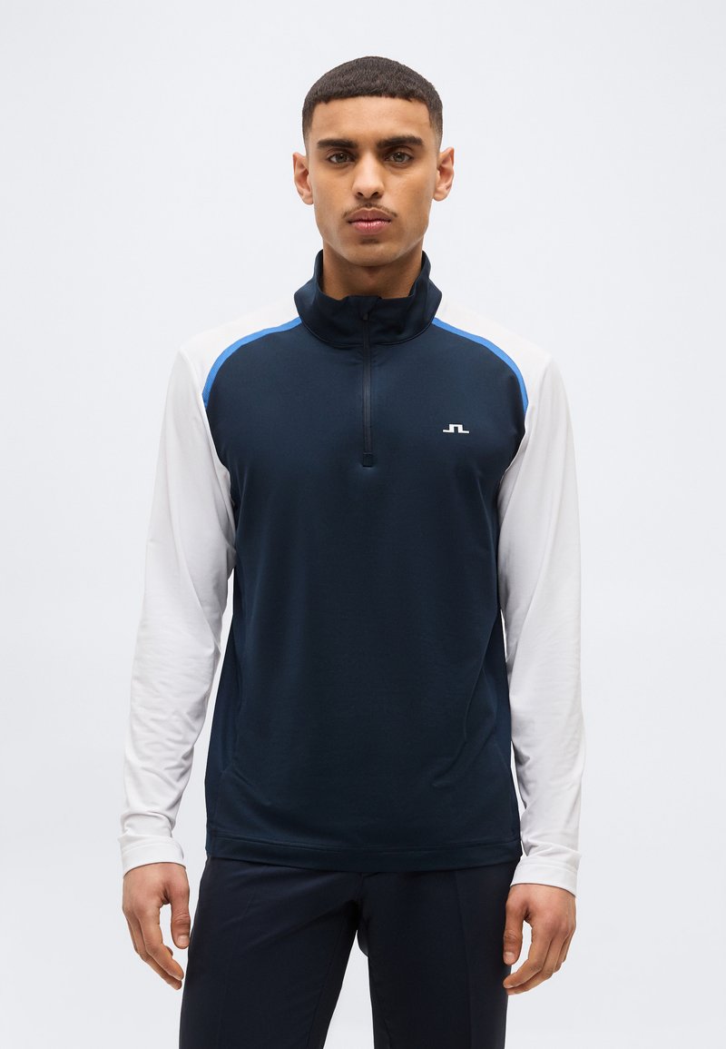 Άνδρας που φοράει αθλητική μπλούζα με φερμουάρ μακρυμάνικη σε ναυτικό μπλε και λευκό χρώμα, με μικρό λευκό λογότυπο στο στήθος, στέκεται μπροστά σε απλό φόντο.