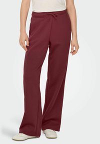 Burgunderfarvede sweatpants med snor i taljen, bred ben-design og blød tekstur, parret med hvide sko.