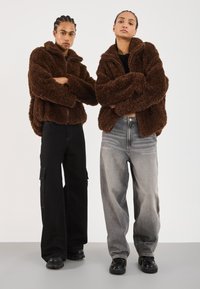 Twee mensen staan met de armen over elkaar, gekleed in bruine fuzzy jassen, brede broeken en zwarte schoenen, tegen een eenvoudige lichte achtergrond.