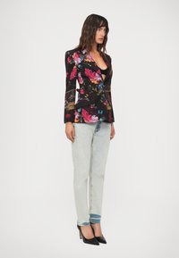 Donna in jeans chiari, tacchi alti neri e un blazer aderente nero con motivi floreali e di uccelli colorati, con capelli scuri stratificati.