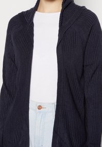 Námořnický žebrovaný cardigan s kapucí, zdobený svislými pruhy a pohodlným střihem. Kombinován s bílým tričkem a světle modrými džíny.