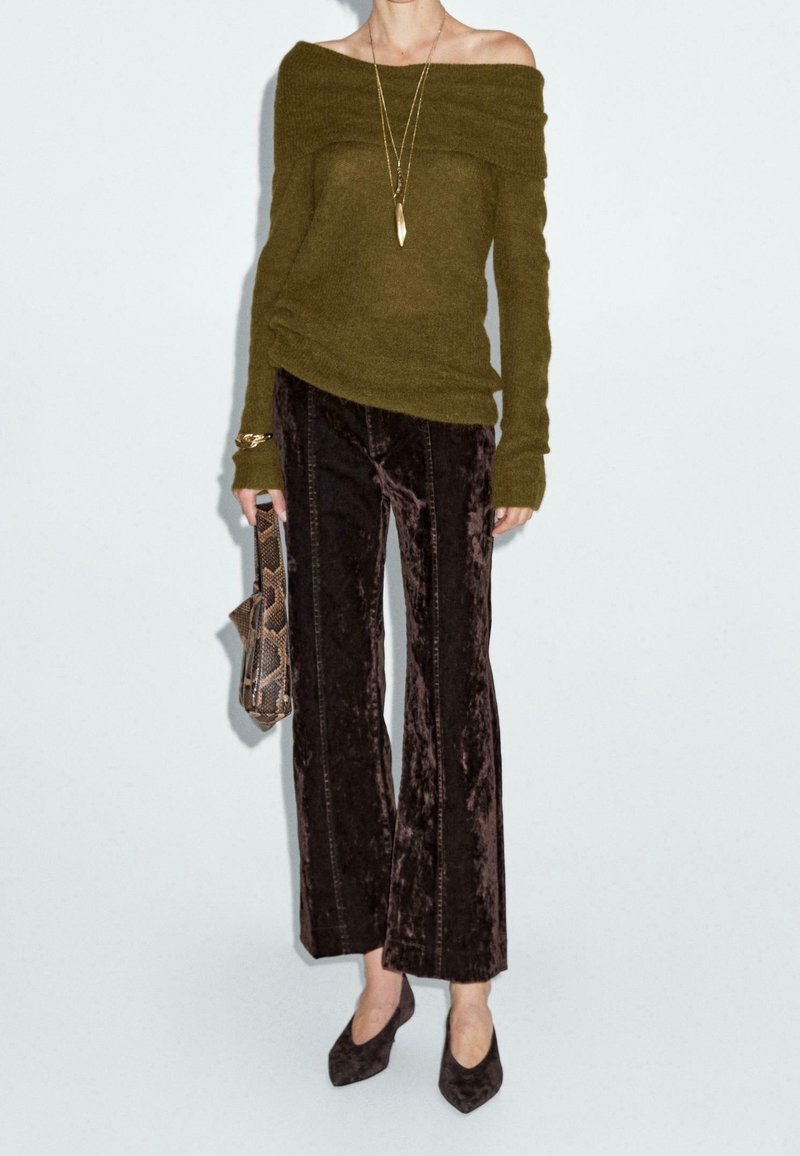 Femme portant un pull vert olive à épaules dénudées, un pantalon évasé en velours marron foncé, des escarpins pointus, un collier en or, et tenant une pochette à motif de peau de serpent.