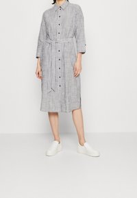 Gestreiftes, knopfgeteiltes Kleid mit einem Taillengürtel, langen Ärmeln und knielangem Saum. Verfügt über schwarze Knöpfe und einen lockeren Schnitt. Weiße Slipschuhe.