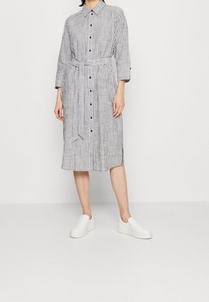 Vestito a righe con bottoni, vita con cintura, maniche lunghe e lunghezza al ginocchio. Presenta bottoni neri e una vestibilità comoda. Scarpe bianche slip-on.