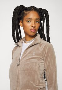 Beige Zip-Jacke aus weichem Samtmaterial mit einem kleinen Logodetail. Das Model hat geflochtenes Haar und trägt ein weißes Oberteil darunter.