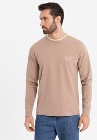 Ein bärtiger Mann trägt ein beiges Langarmshirt mit einem kleinen "EX"-Logo auf der Brust und blaue Jeans. Er schaut nach links.
