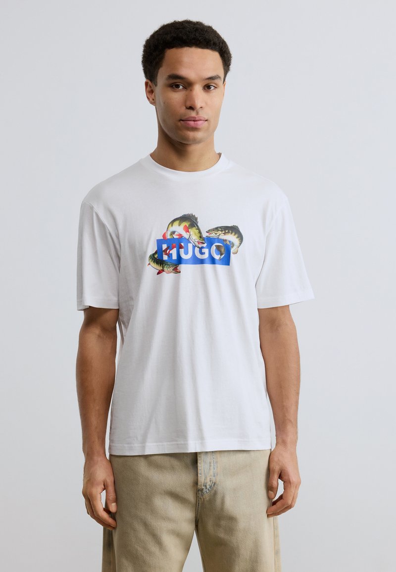 Νεαρός άνδρας που φοράει λευκό T-shirt με πολύχρωμα σχέδια ψαριών και το κείμενο «HUGO», συνδυασμένο με ανοιχτόχρωμο μπεζ παντελόνι, πάνω σε απλό φόντο.