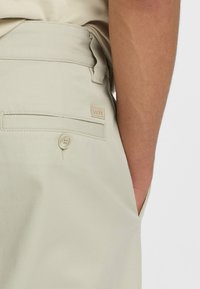 Pantaloni beige su misura con una texture liscia, caratterizzati da una tasca posteriore con chiusura a bottone e un piccolo etichetta del marchio sulla vita.