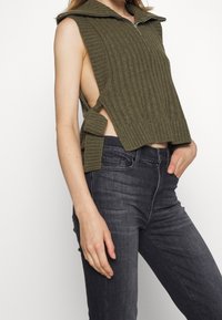 Haut sans manches côtelé vert olive avec un col zippé, présentant des découpes latérales et un détail noué à la taille. Associé à un jean foncé.