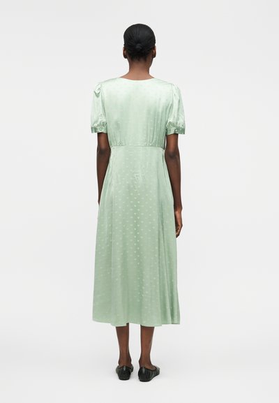 Γυναίκα με σκουρόχρωμη επιδερμίδα και μαλλιά σε κότσο, που φοράει ένα ανοιχτοπράσινο φόρεμα μέχρι το γόνατο με φουσκωτά μανίκια και μαύρα χαμηλά παπούτσια, στέκεται γυρισμένη με την πλάτη.