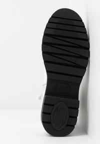 Semelle en caoutchouc noire texturée d'une botte avec des rainures profondes et un logo ovale, positionnée verticalement sur un fond blanc.