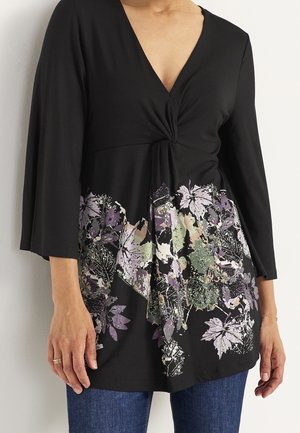 Sort V-halsbluse med snoet front og bladmønster i lilla, grøn og beige ved taljen, båret med blå jeans og guldsmykker.