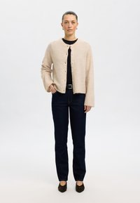 Beige stickad cardigan med rund halsringning och knappstängning, kombinerad med mörkblå denimjeans och mörkbruna skor.