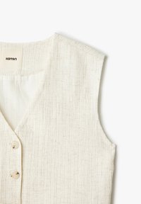 Cremefarbener ärmelloser Weste mit zwei Knöpfen und V-Ausschnitt, der einen teilweise sichtbaren Markenaufnäher im Kragen zeigt.