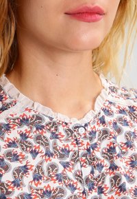 Hvid bluse med et paisley-blomstermønster i blå, rød og brun. Har en flæsekant ved halsen og en knaplukning foran.