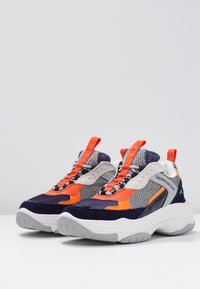 Pár robustních tenisek v námořnické modré, oranžové, šedé a bílé barvě s akcenty z síťoviny a semiše, vybavený jasně oranžovými poutkami a brandingem Calvin Klein Jeans.